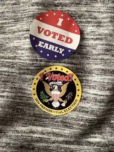 Two circular U.S. election pins are pinned to a gray and white heathered fabric. The top pin is red, white, and blue with…