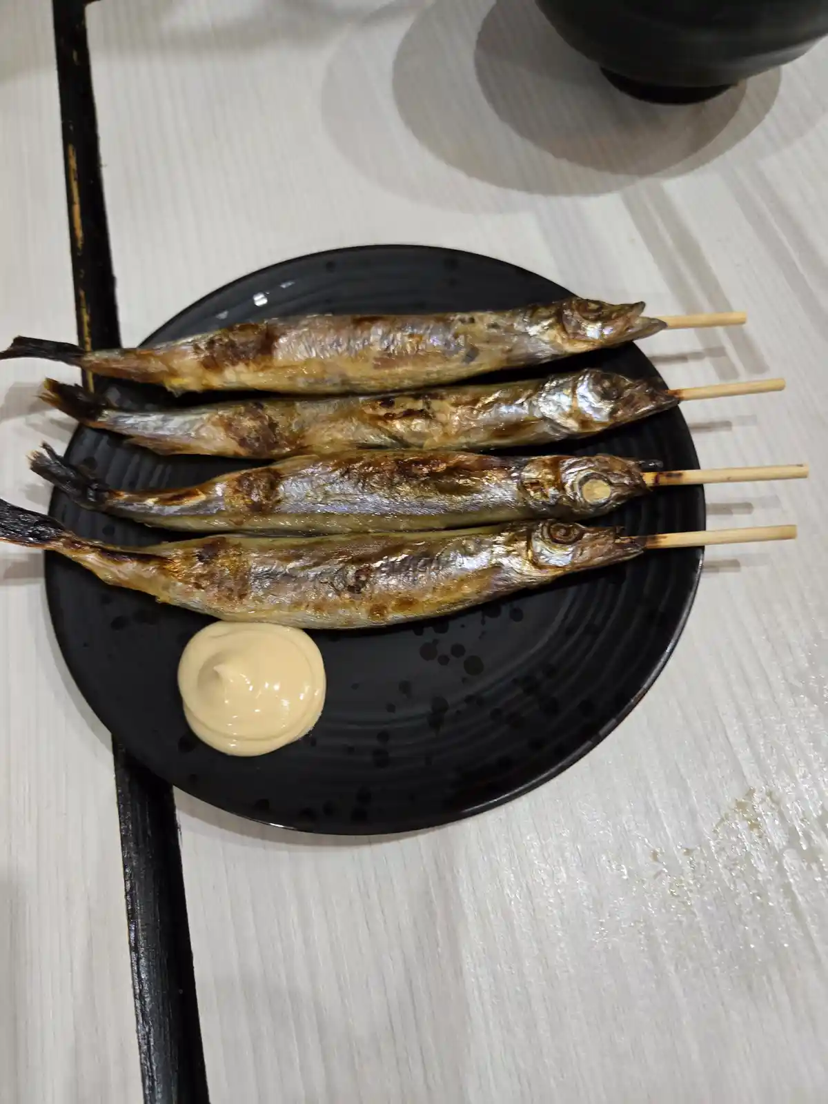 A black plate holds four grilled whole fish on wooden skewers, arranged neatly. A dollop of creamy yellow sauce sits beside…