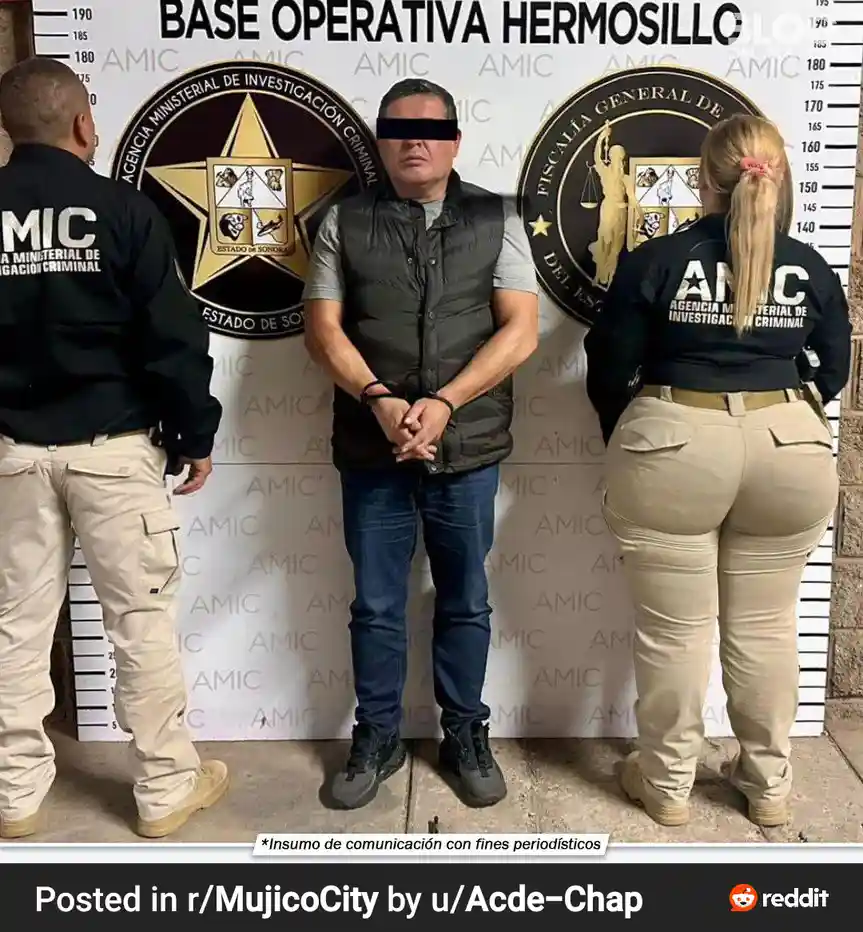 A man in a vest and jeans stands with hands cuffed in front of him, flanked by two uniformed officers in black shirts and…