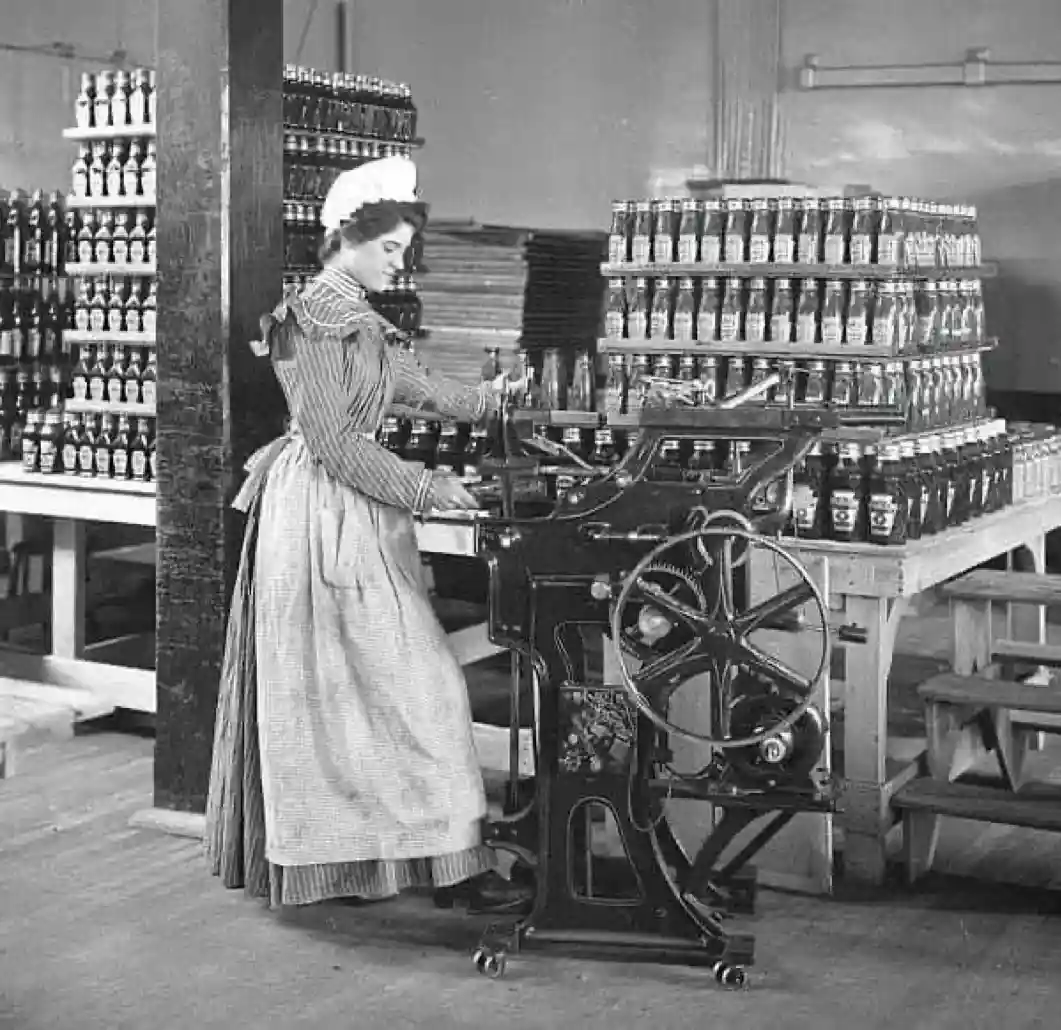 A vintage black-and-white photo shows a woman in a long dress and bonnet operating an old-fashioned bottling machine in a…