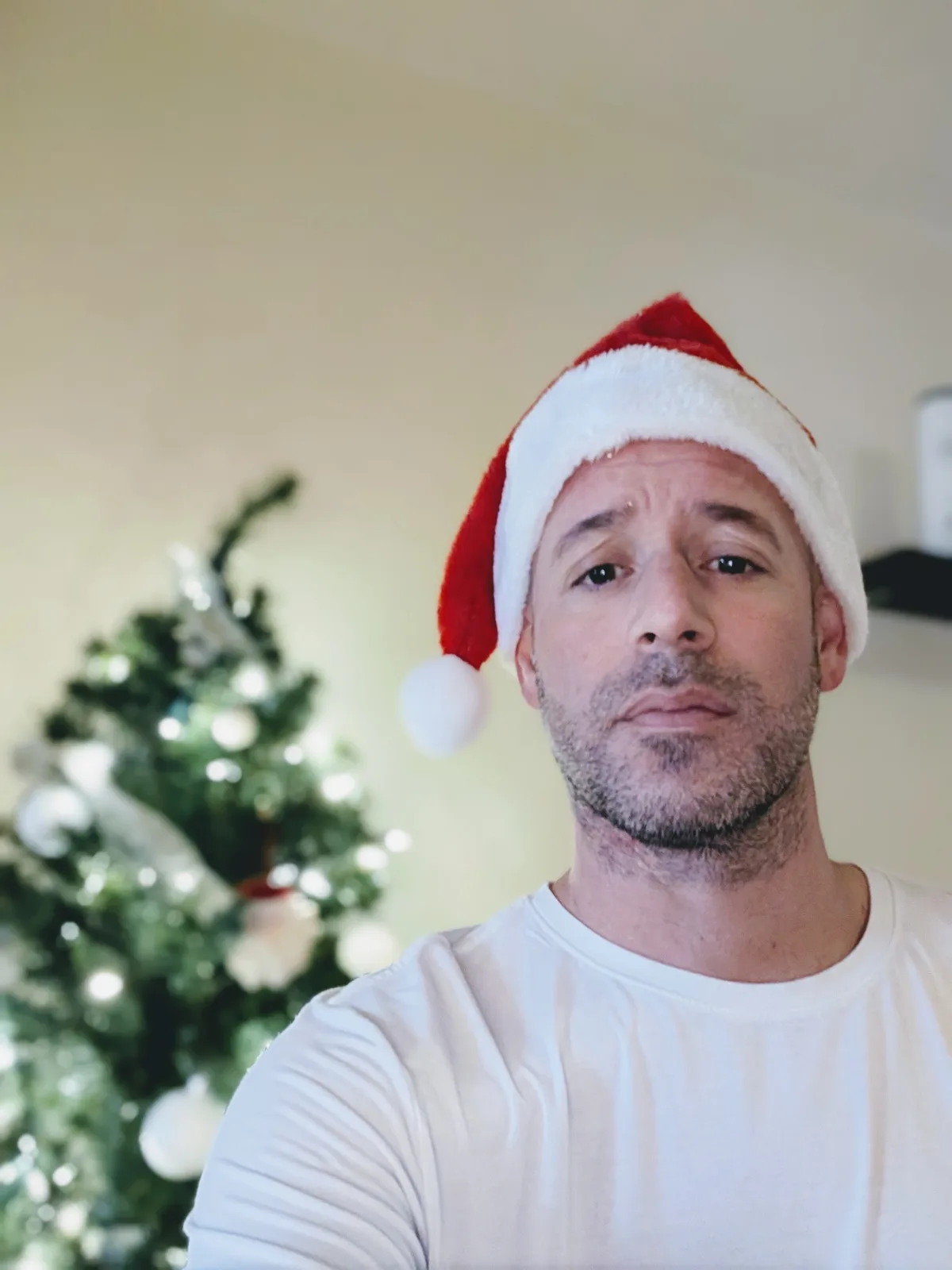A man wearing a red and white Santa hat looks directly at the camera with a weary, unenthusiastic expression. Behind him, a…