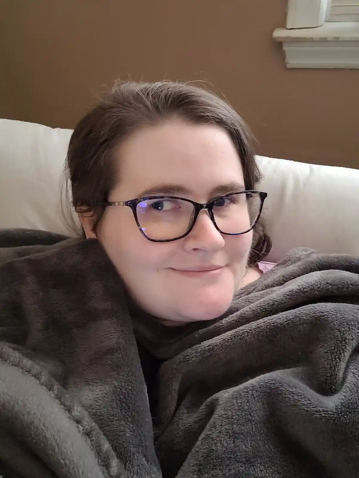 A person with glasses and brown hair is wrapped in a dark grey blanket while sitting on a white couch, looking slightly to…