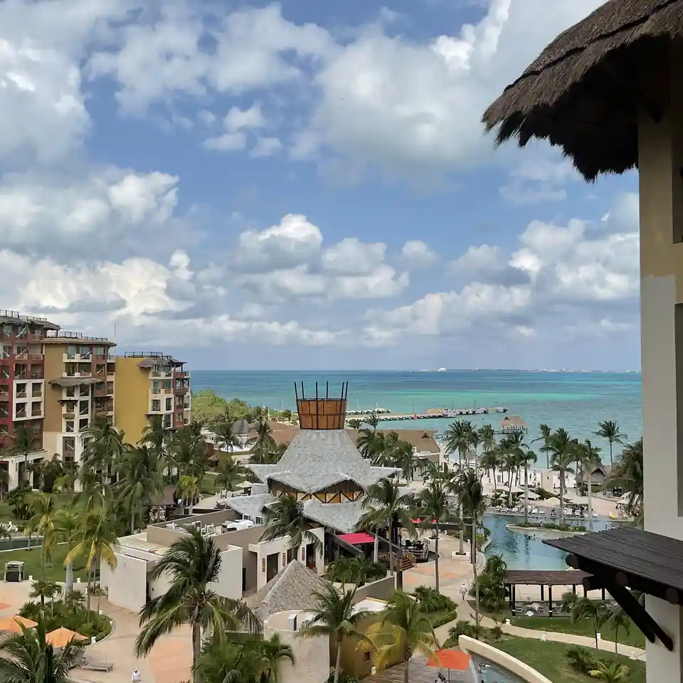 A scenic view from a resort balcony overlooking turquoise ocean water, palm trees, and multi-story buildings with thatched…