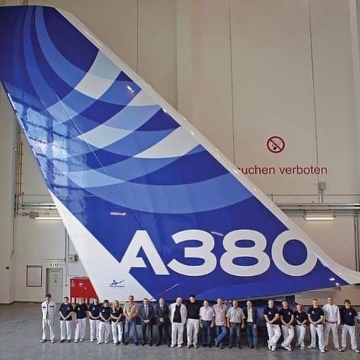 A group of people stands in front of the massive tail fin of an Airbus A380 airplane inside a hangar. The tail fin is blue…