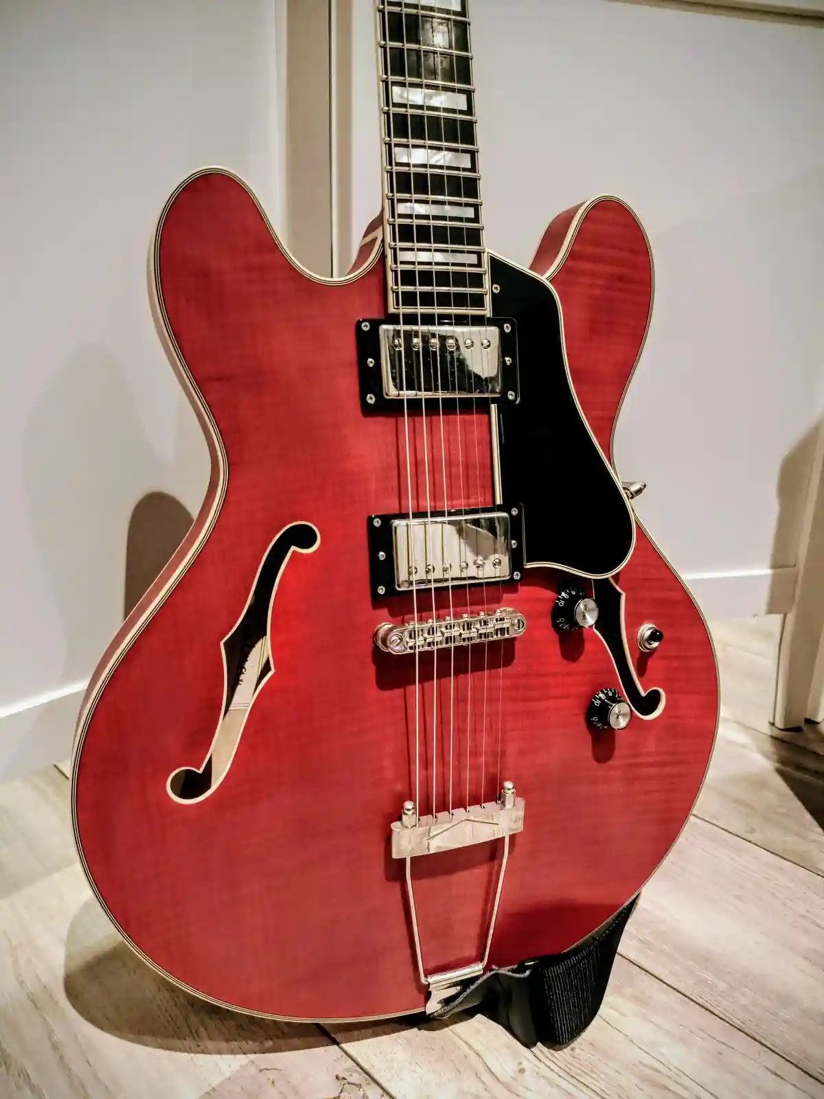 A red semi-hollow body electric guitar with f-holes, chrome hardware, and a black pickguard stands upright on a light wood…