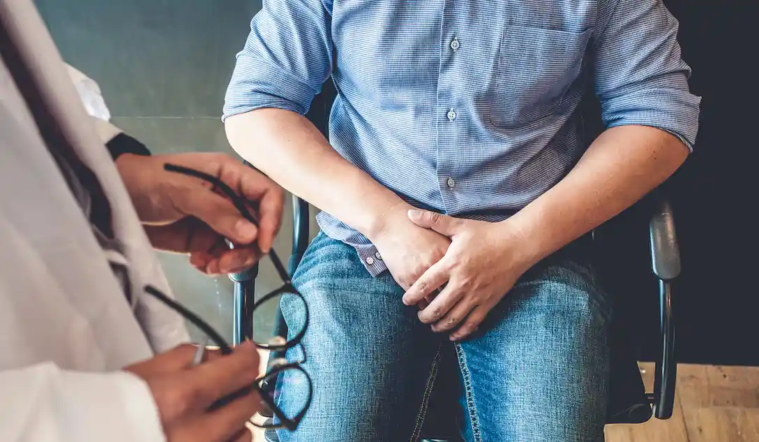 A man in a blue checkered shirt and jeans sits in a chair with both hands clasped over his lower abdomen, appearing anxious…