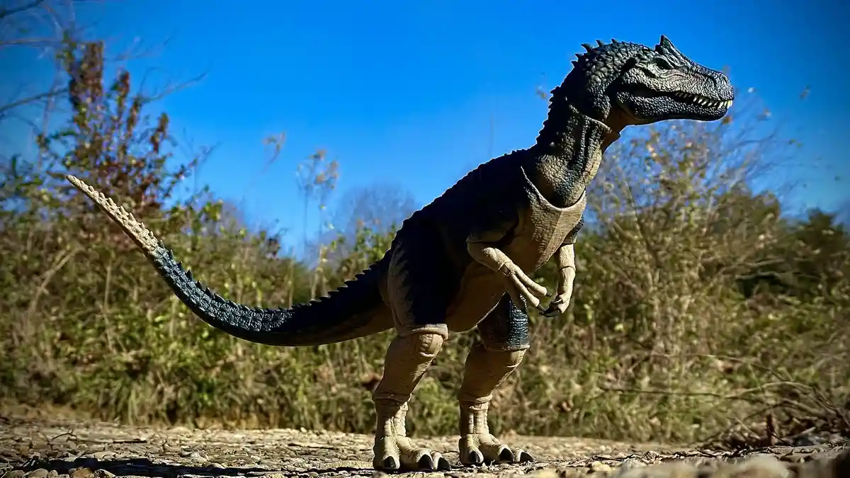 A toy dinosaur figure, resembling a theropod with dark blue and beige coloring, stands outdoors on gravel with bushes and a…