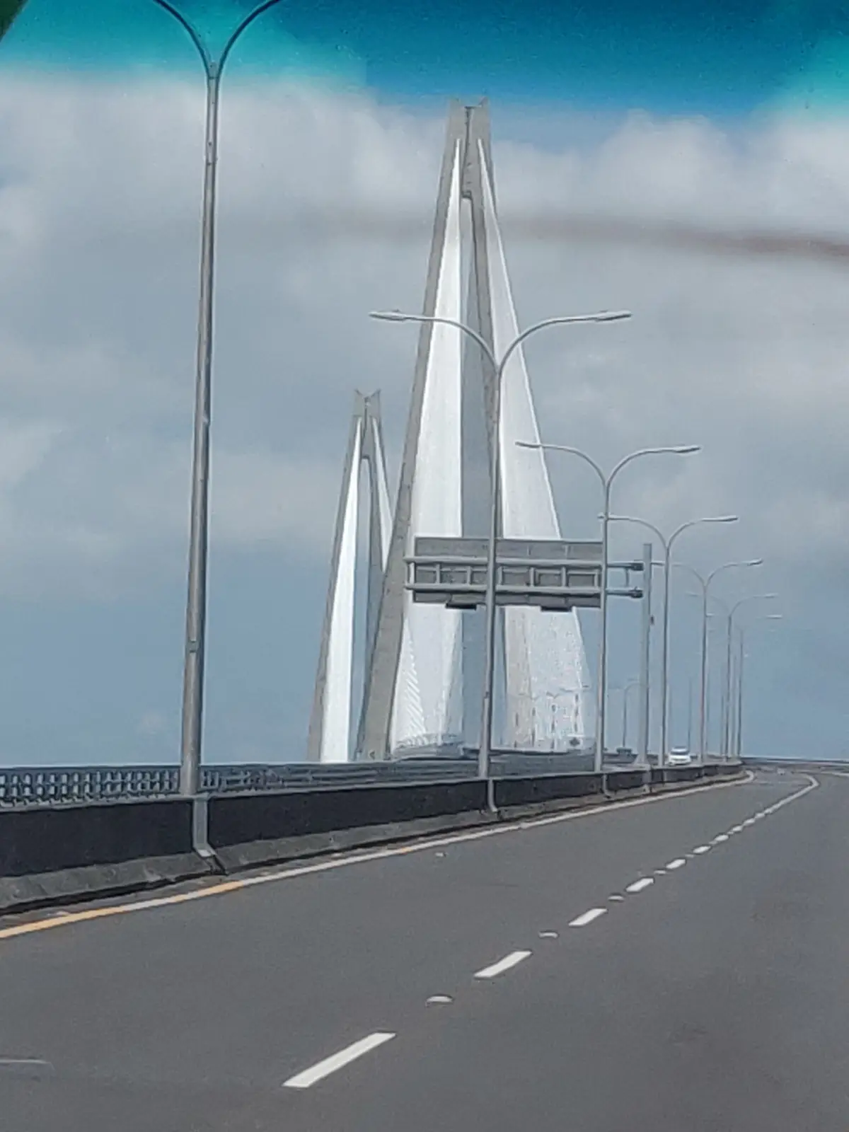 A photo taken from a car on a highway shows a massive, modern cable-stayed bridge with tall, white, V-shaped pylons…