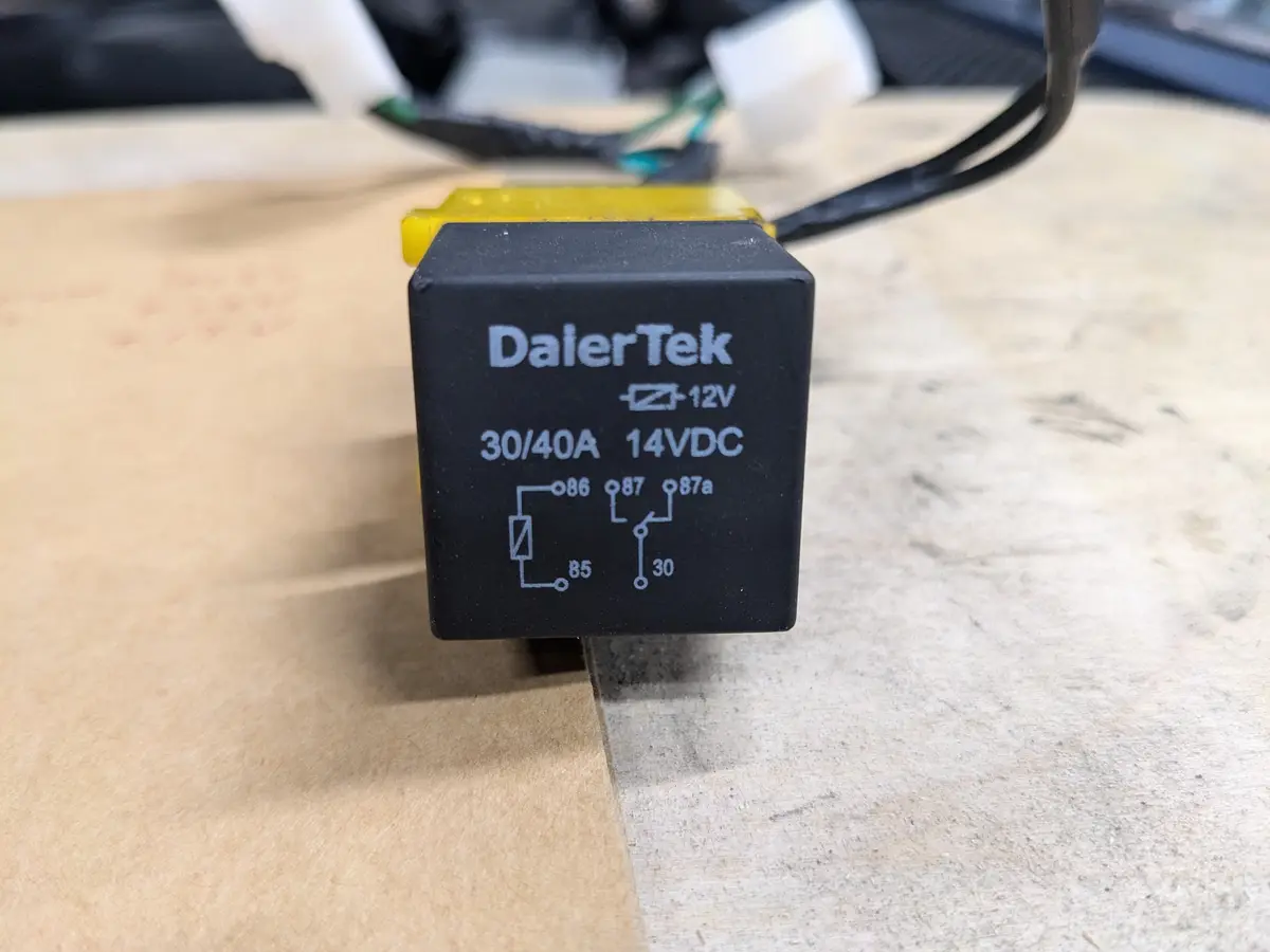 A close-up photo of a black DaierTek automotive relay with white printed text and pin diagram, sitting on a workbench next…