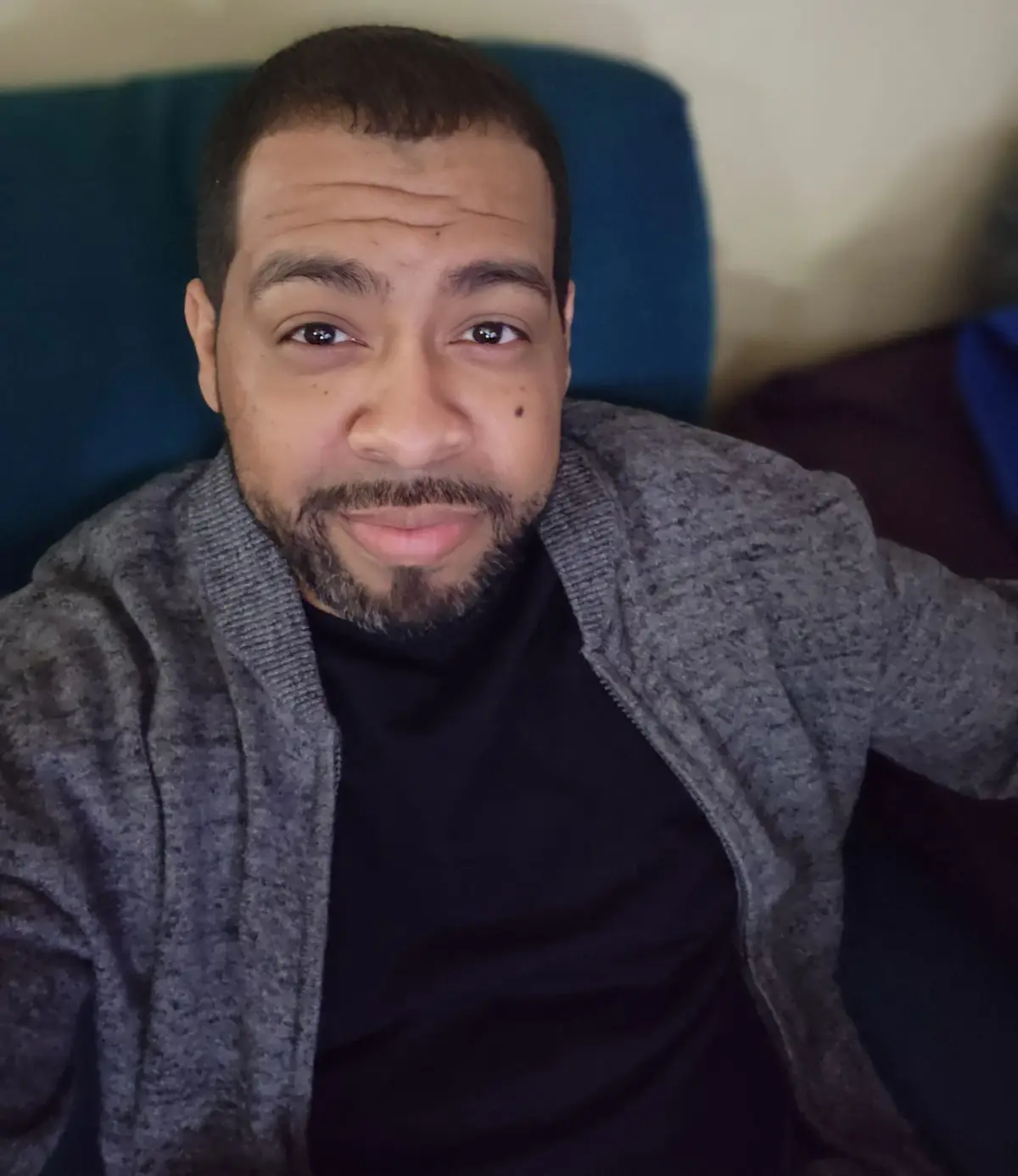 A man with short dark hair and a beard, wearing a grey cardigan over a black shirt, sits on a blue couch and looks directly…