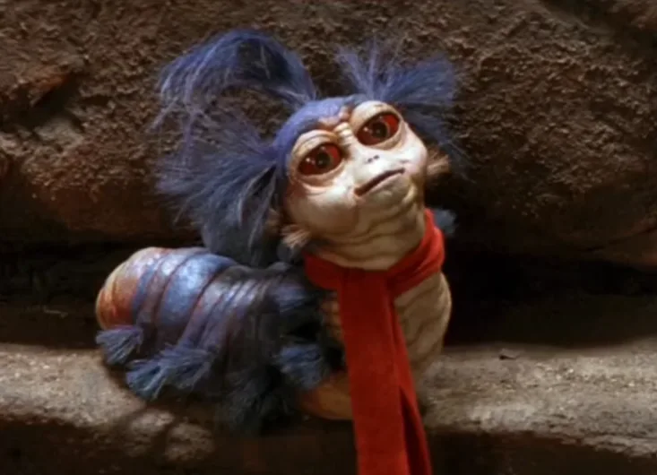 A close-up of a small, furry alien creature with large red eyes and blue hair, wearing a red scarf, looking upward with a…