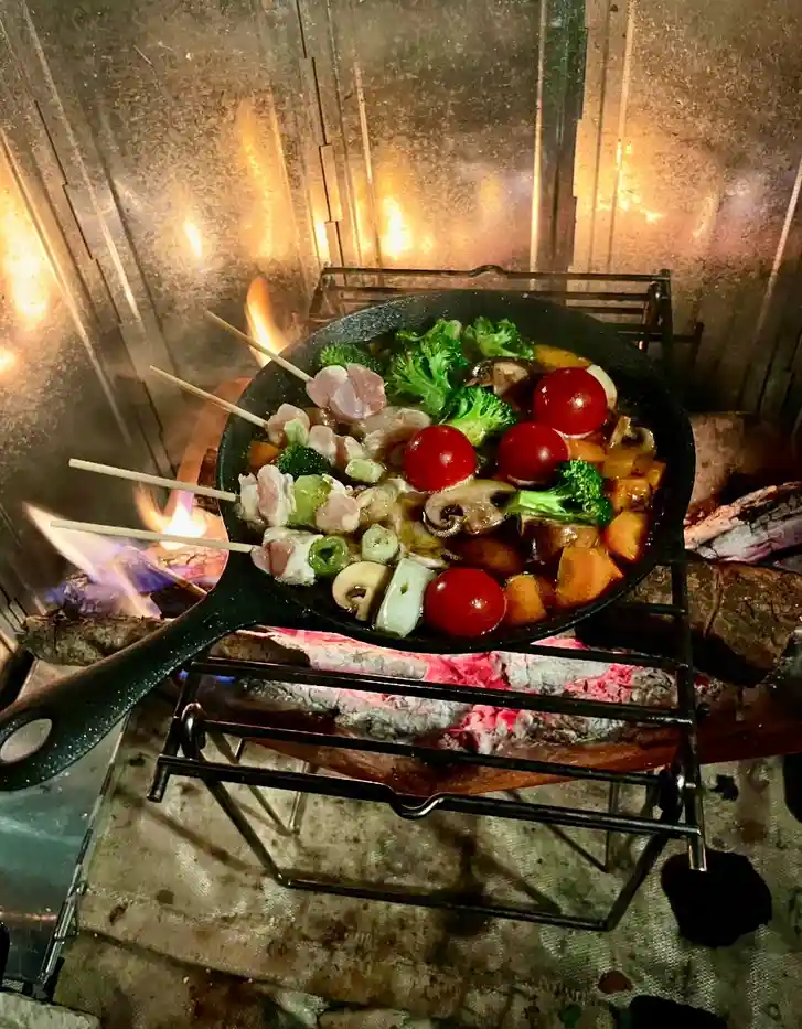 A black cast iron skillet filled with colorful vegetables, cherry tomatoes, mushrooms, and meat pieces on skewers is placed…