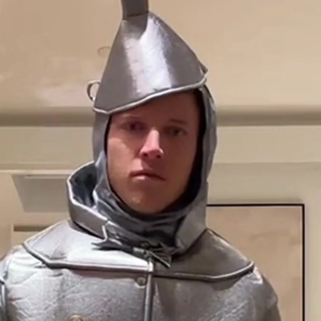 A man wearing a full silver tin man costume from The Wizard of Oz, including a pointed helmet and hood, stares blankly at…