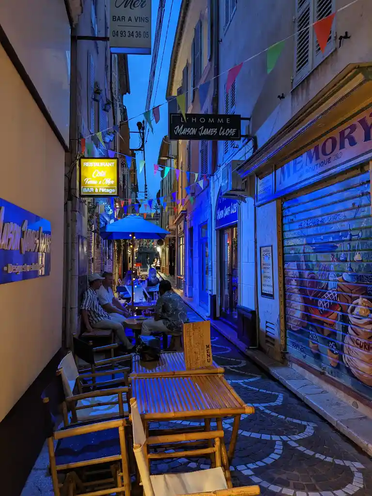 A narrow, cobblestoned European alleyway at dusk, illuminated by blue ambient lighting and string flags. People sit at…
