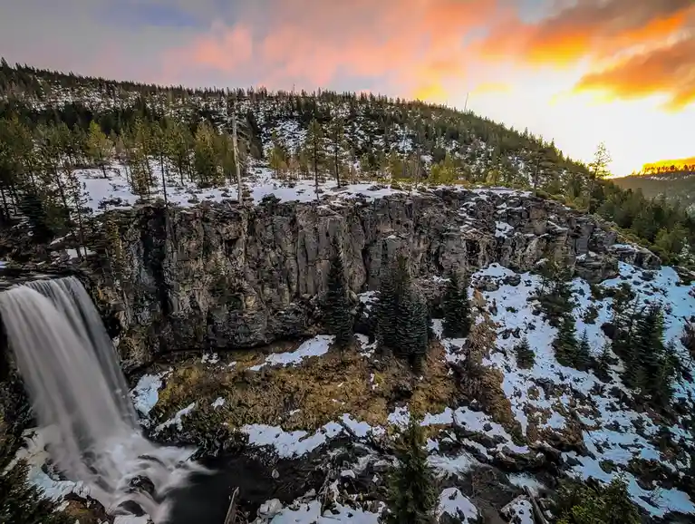A scenic winter landscape featuring a waterfall cascading over a rocky cliff, surrounded by snow-dusted evergreen trees…