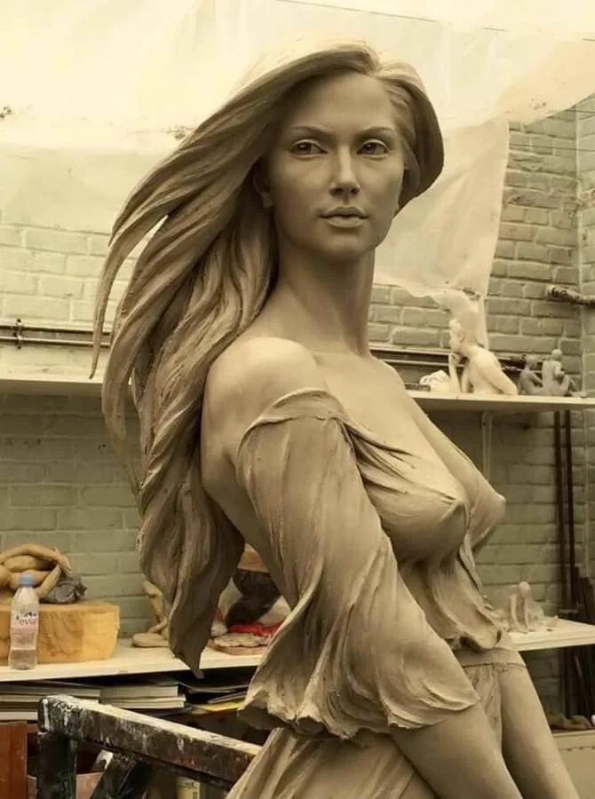 A highly detailed, life-sized clay sculpture of a woman with long flowing hair and a draped garment, posed in a studio with…