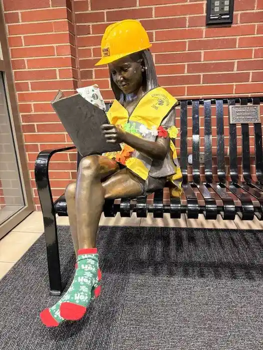 A bronze statue of a young girl sitting on a black metal bench, wearing a yellow hard hat, a yellow safety vest, and festive…