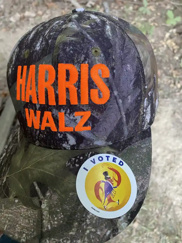 A close-up of a camouflage baseball cap with bright orange embroidered text reading 'HARRIS WALZ' on the front. Attached to…