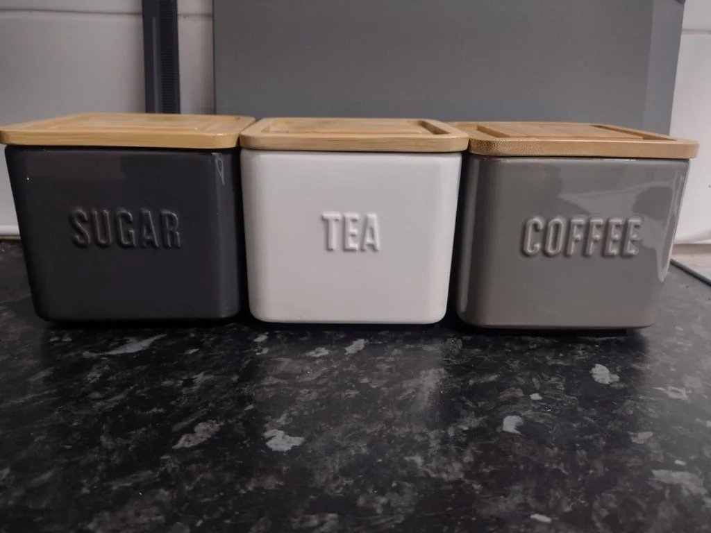 Three ceramic kitchen canisters with bamboo lids sit side-by-side on a dark countertop; the left is black and labeled…