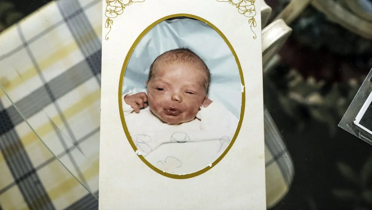 A vintage-style baby photo of a newborn with a slightly chubby face and closed eyes, framed in an oval with gold trim on a…