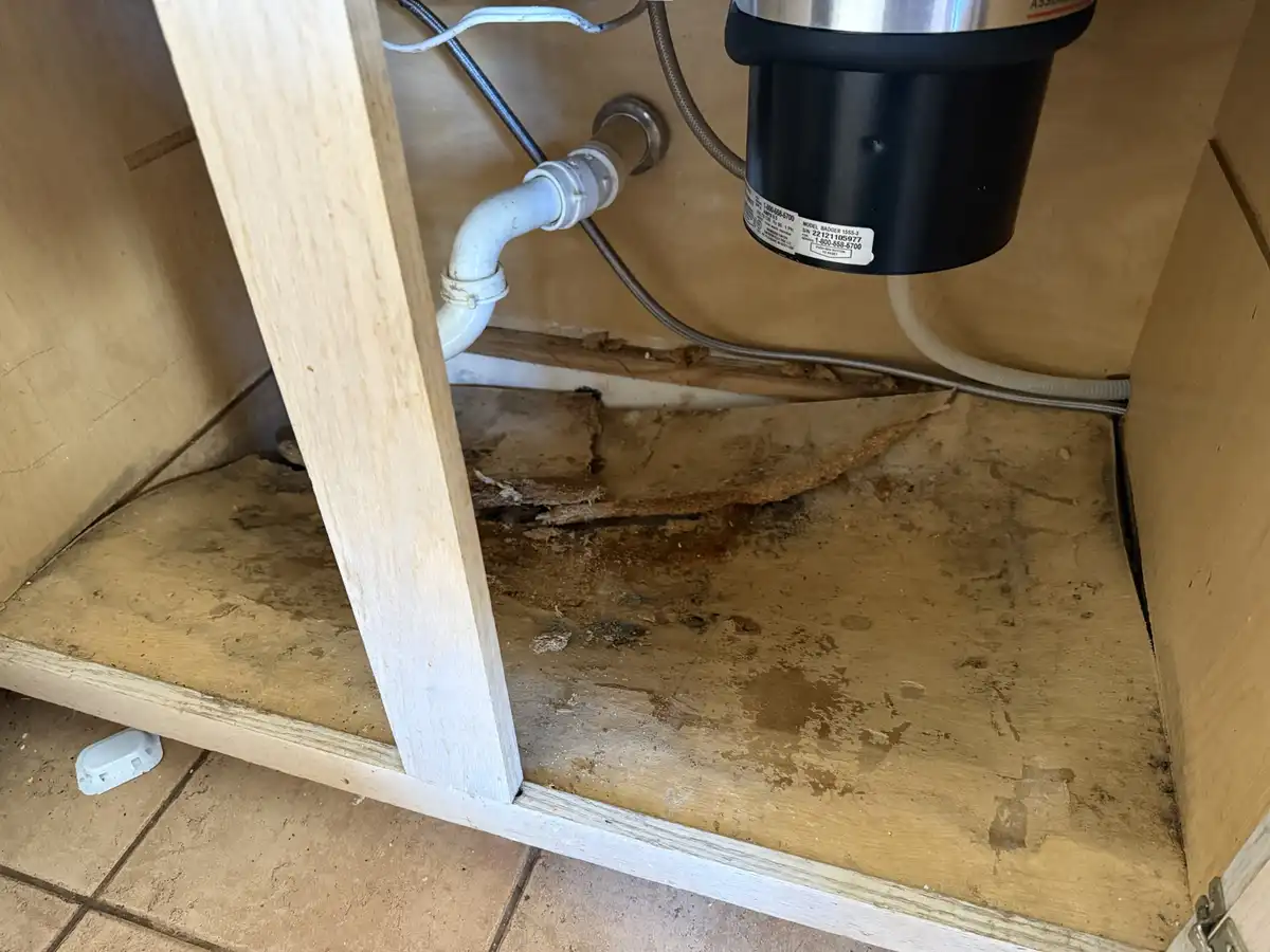 A close-up photo shows the underside of a kitchen cabinet with visibly damaged, water-stained, and rotting plywood flooring…