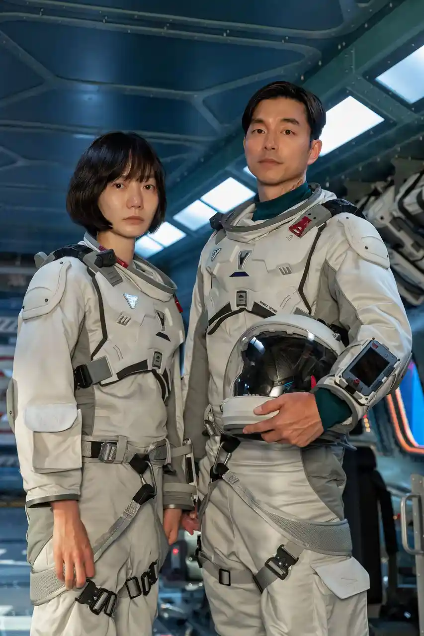 Two people in detailed white astronaut suits stand inside a futuristic spacecraft, looking serious. The man on the right…
