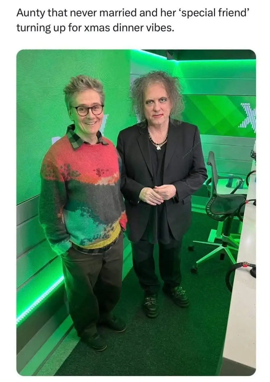 Two women stand side by side in a green-lit studio. The woman on the left wears glasses and a colorful sweater, smiling…