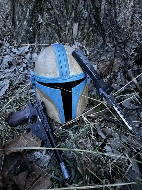 A blue and tan Mandalorian helmet lies on the ground among dry leaves and grass, with a blaster pistol and a knife resting…