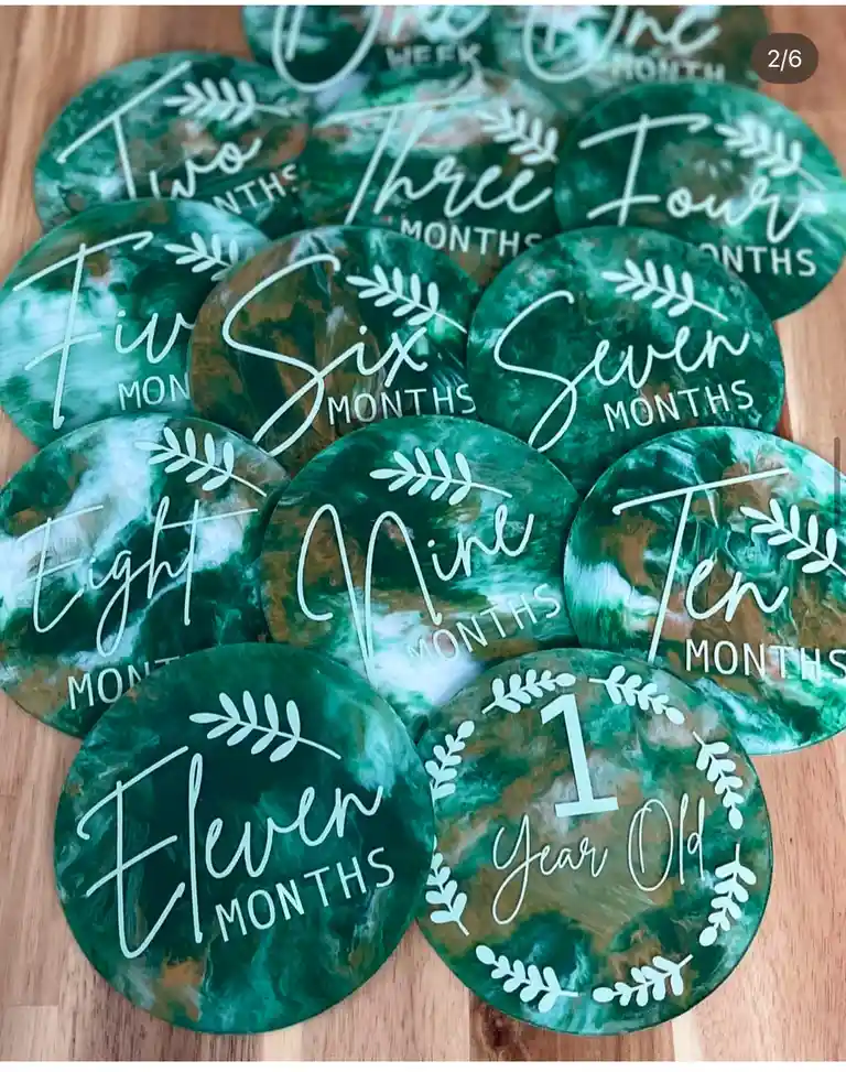 A collection of green marbled circular milestone markers for a baby, each engraved with a month or age milestone from 'One…