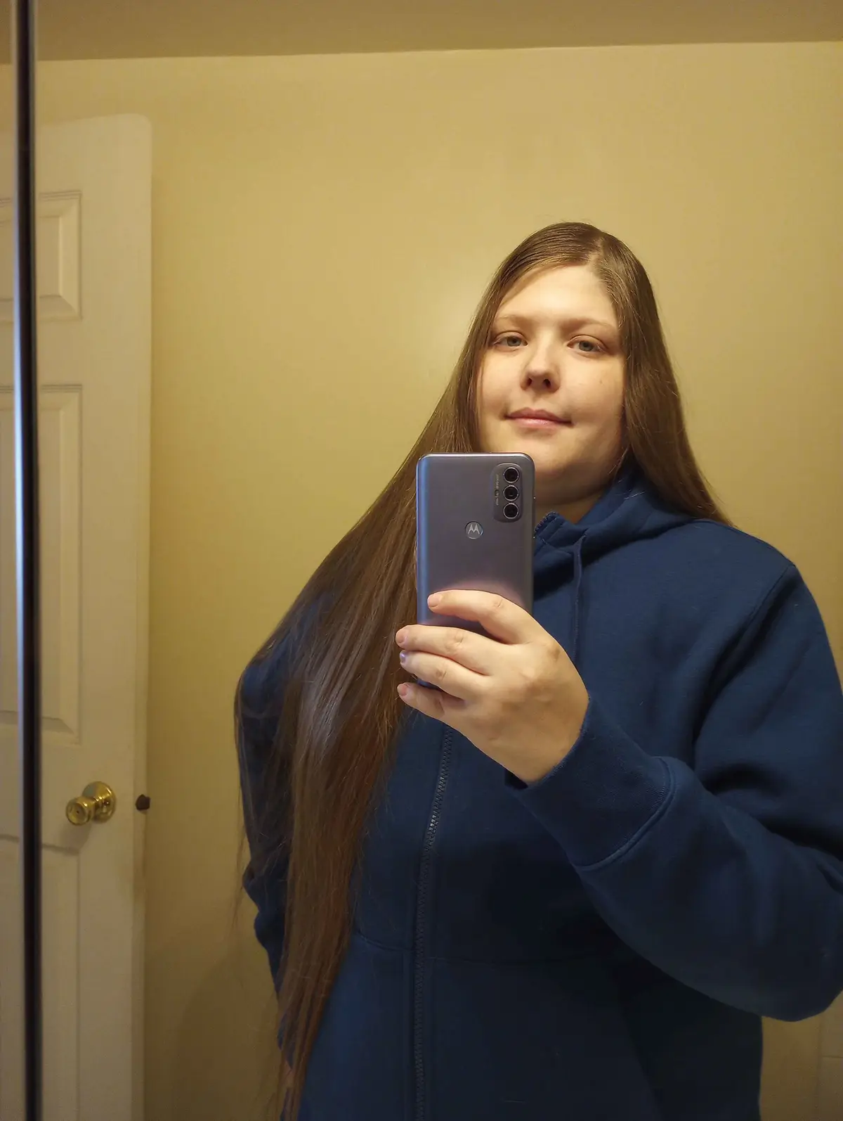 A person with long brown hair wearing a blue zip-up hoodie takes a mirror selfie with a gray Motorola phone, smiling…