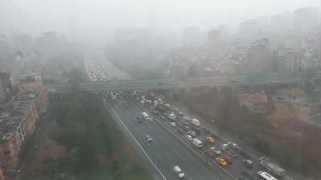 An aerial view of a city highway during heavy rain and fog, showing multiple lanes of congested traffic with cars and trucks…