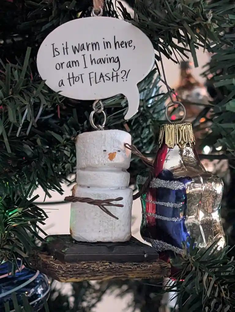 A close-up of a Christmas tree ornament featuring a snowman made of three stacked marshmallows with twig arms and a carrot…
