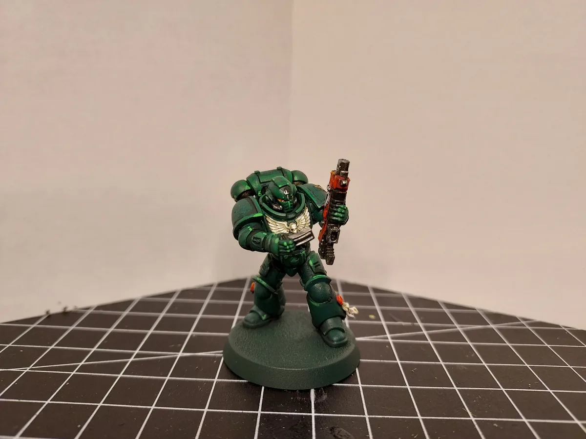 A painted green Space Marine miniature in power armor stands on a circular base atop a black grid-patterned surface, holding…
