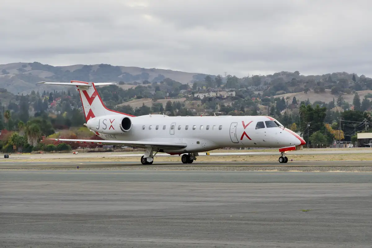 A white private jet with red 'X' logos on its tail, fuselage, and nose is taxiing on an airport runway. The aircraft's…