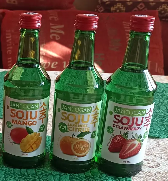Three green glass bottles of Jantugan soju, each with a different fruit flavor label—Mango, Japanese Citrus, and…