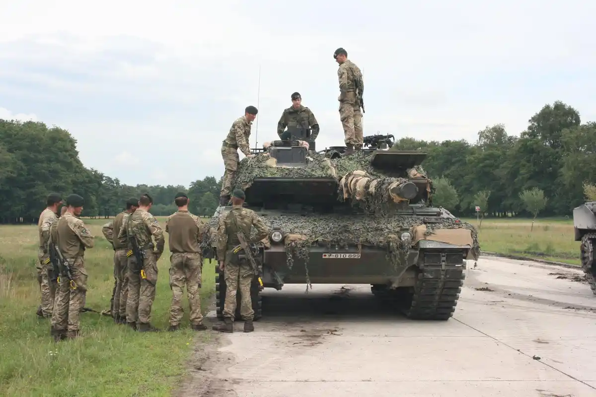 A group of soldiers in camouflage uniforms stand around a camouflaged military tank on a paved road in a grassy field. Three…