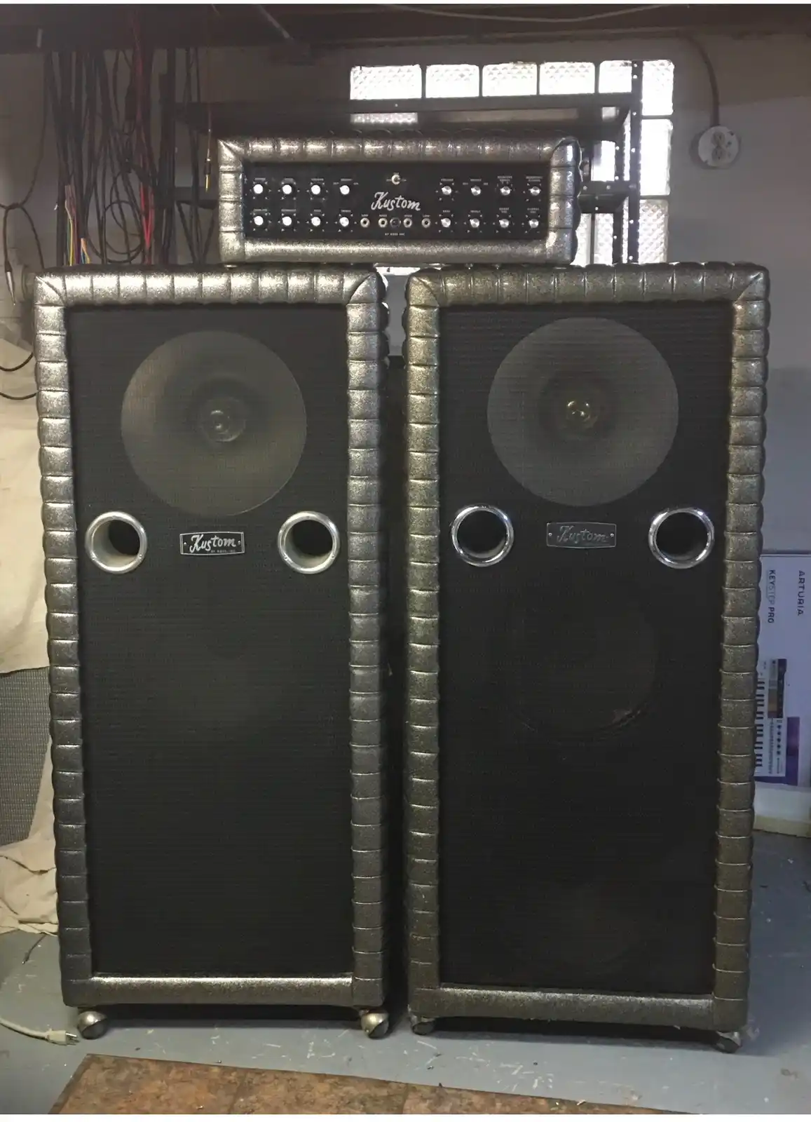 Two large, silver-framed Kustom guitar amplifiers with black speaker grilles and circular ports are stacked side-by-side in…