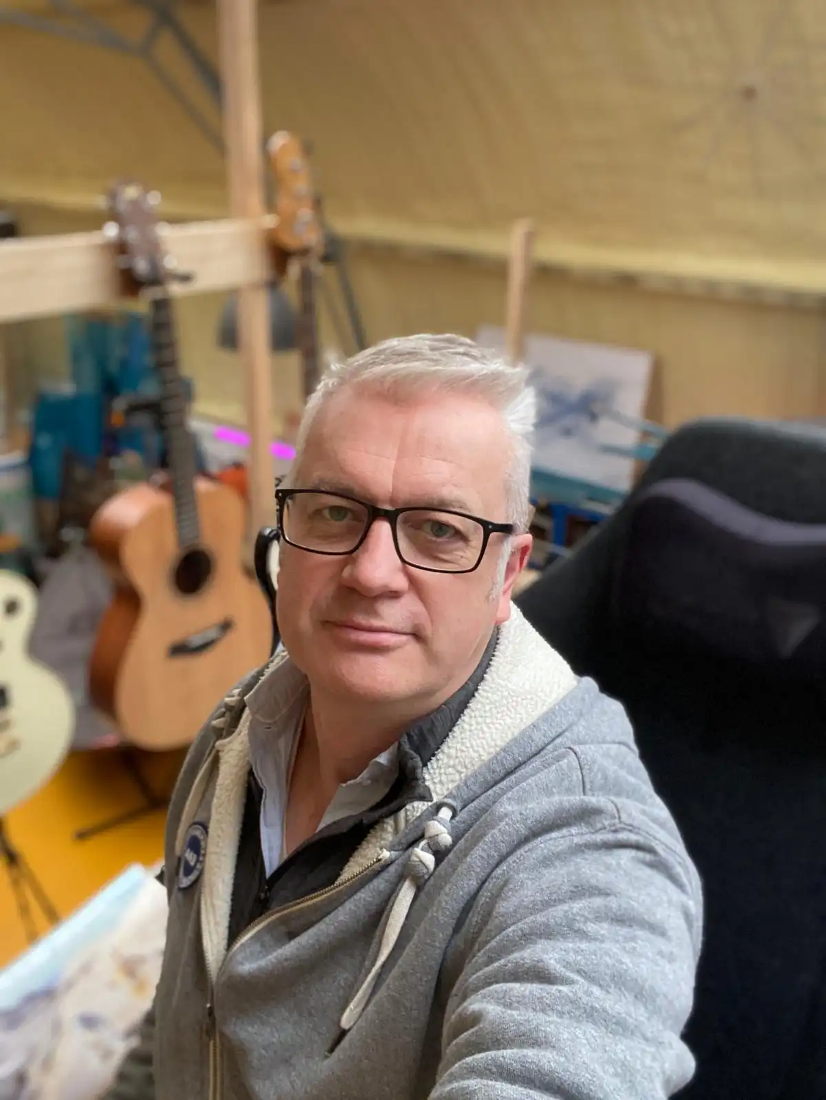 A middle-aged man with glasses and gray hair takes a selfie in a room filled with guitars and musical equipment. He wears a…