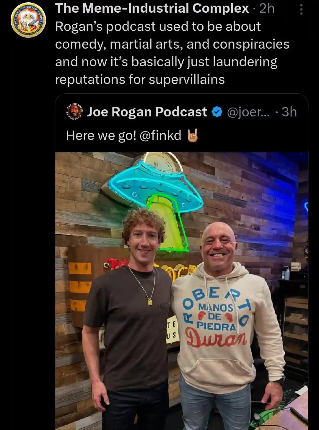 Two men, Mark Zuckerberg and Joe Rogan, are smiling and posing for a photo in front of a large, glowing blue and green…