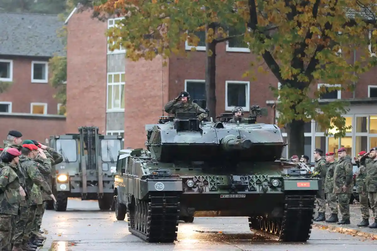 A German military tank, marked with the number 70 and registration Y-810 907, drives down a wet street past rows of soldiers…