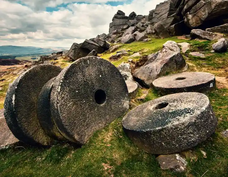 Several large, weathered stone millstones lie scattered on a grassy hillside with rocky outcrops and a cloudy sky in the…
