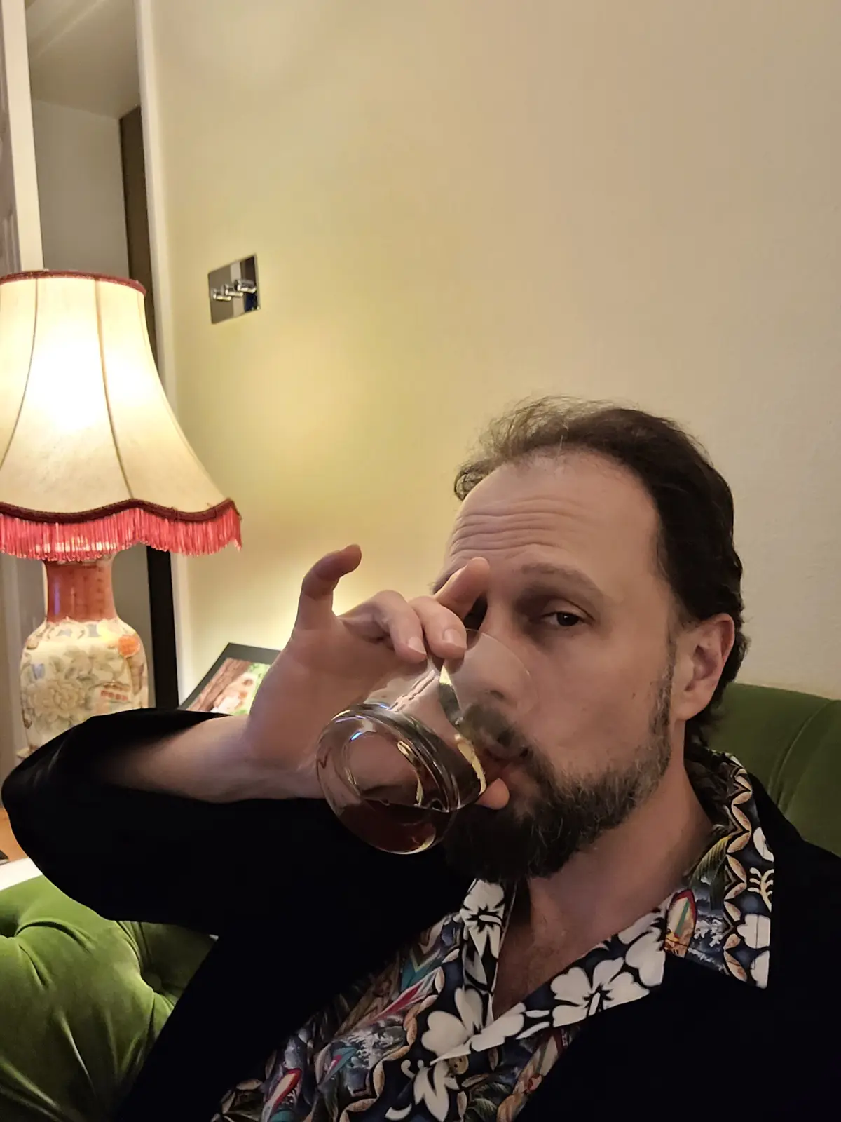 A man with a beard and a patterned shirt is sitting on a green couch, holding a glass of amber liquid to his lips as if…