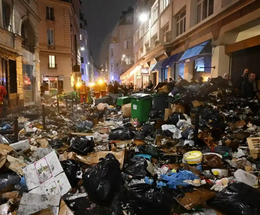 A nighttime street scene shows a massive, chaotic pile of garbage bags, cardboard, and debris filling the road between…