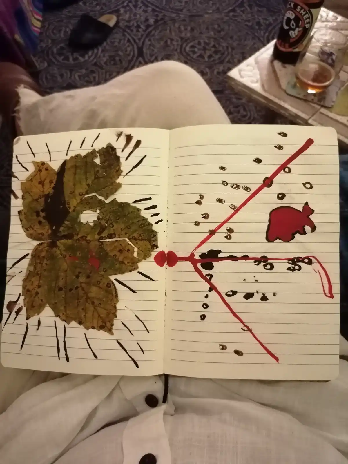 An open lined notebook rests on someone's lap, showing a half-finished art project. The left page features a pressed leaf…