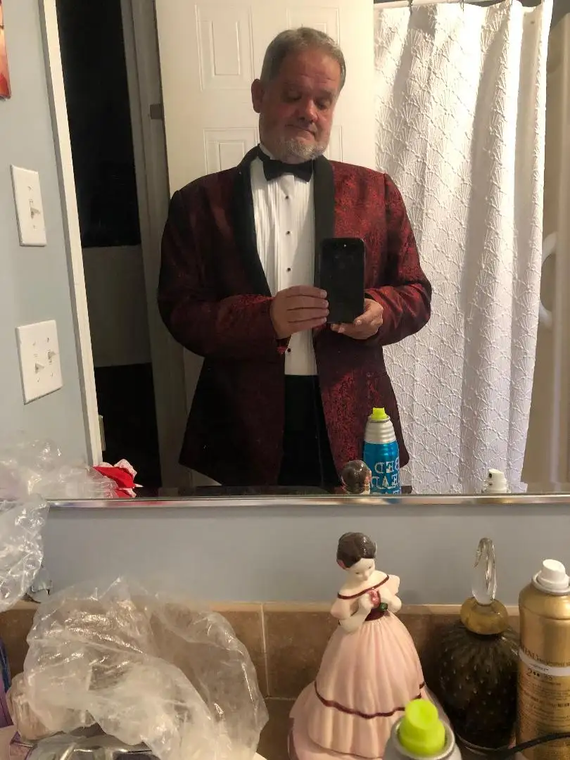 A man in a red patterned tuxedo jacket and black bowtie takes a mirror selfie in a bathroom, holding his phone. The bathroom…