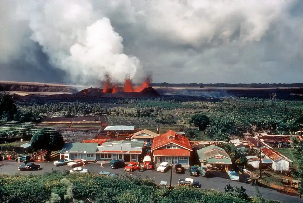 A vintage photograph shows a small town with a gas station, parked cars, and buildings in the foreground, while a large…