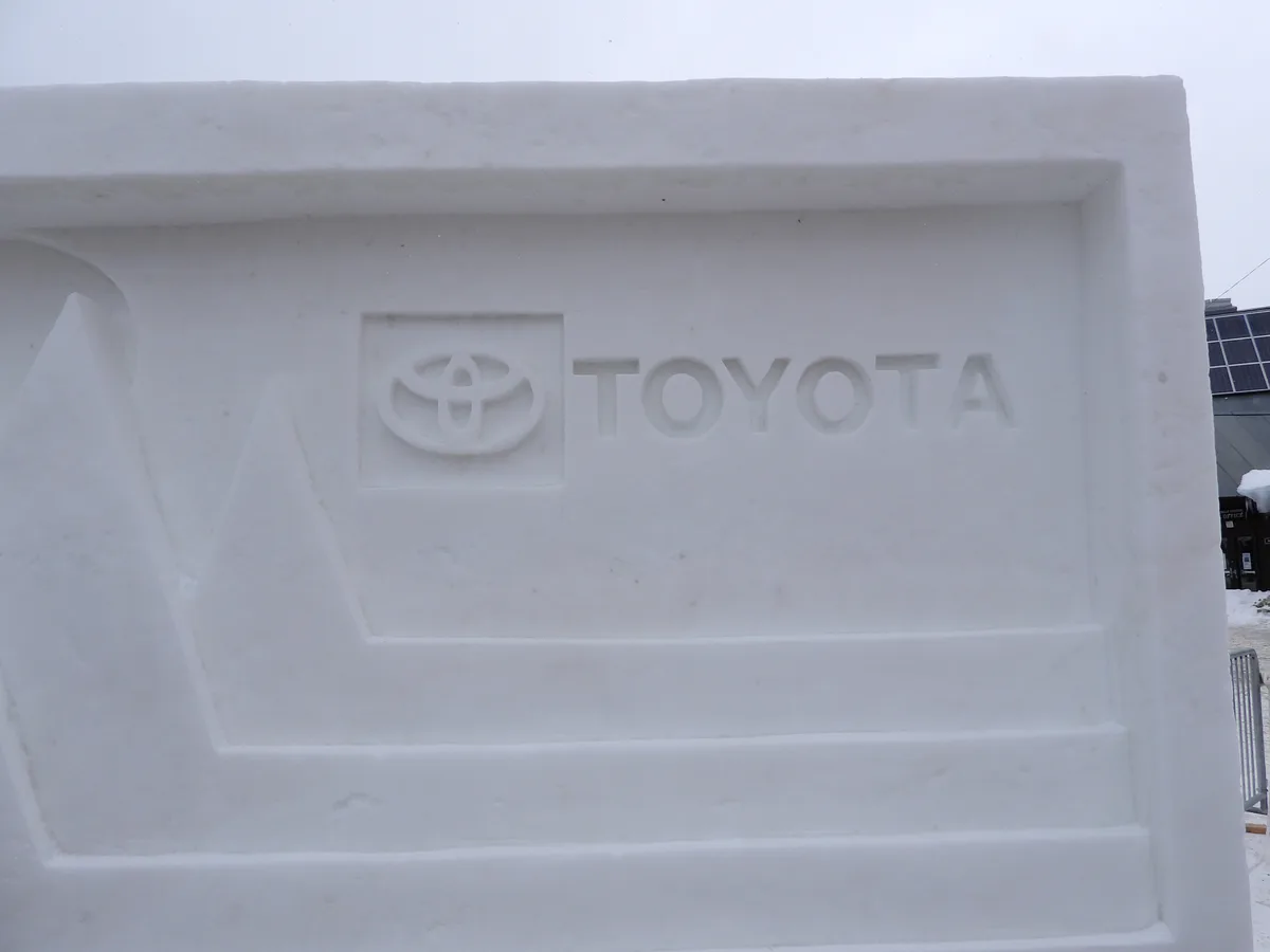 A close-up of a snow sculpture carved to resemble a Toyota car emblem and brand name, with the iconic three-oval logo and…