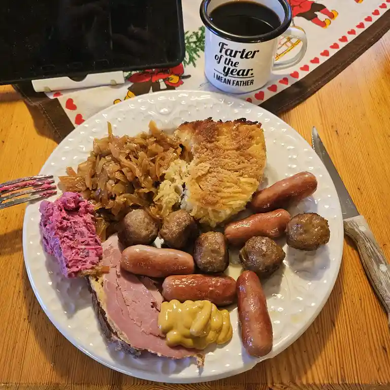 A plate of heavy breakfast food including sausages, meatballs, ham, potato pancake, sauerkraut, and pink beet salad sits on…