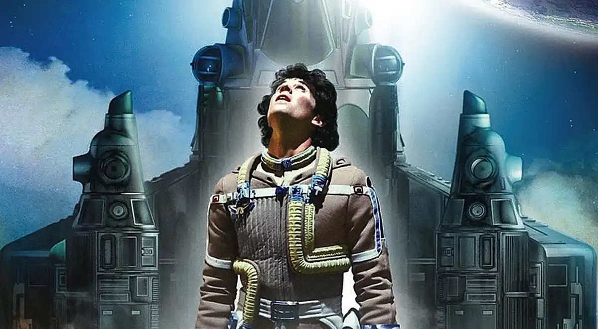 A man in a brown flight suit with yellow and blue straps looks upward in awe against a backdrop of large, futuristic…
