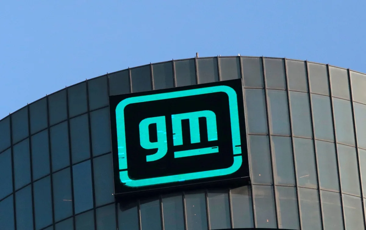 A large, glowing teal GM logo is mounted on the curved glass facade of a modern building against a clear blue sky. The logo…